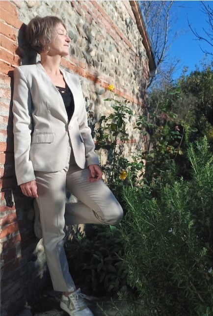 Katja Rehse standing sideways beside a stone wall in a garden, eyes closed, face turned towards the sun with a gentle smile.