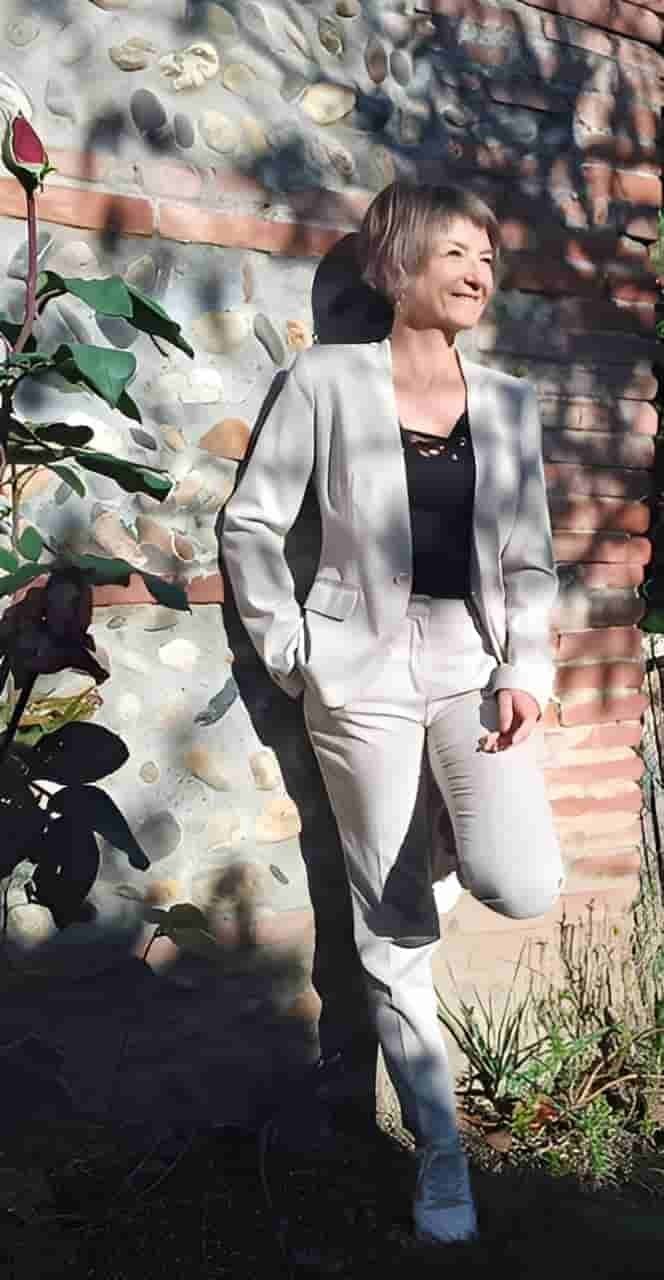 Katja Rehse standing beside a stone wall, one leg resting against it, looking towards the sun next to a rose bush.