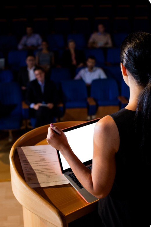 A dark-haired woman seen from behind, holding a laptop, addresses an audience from a wooden lectern in an auditorium.