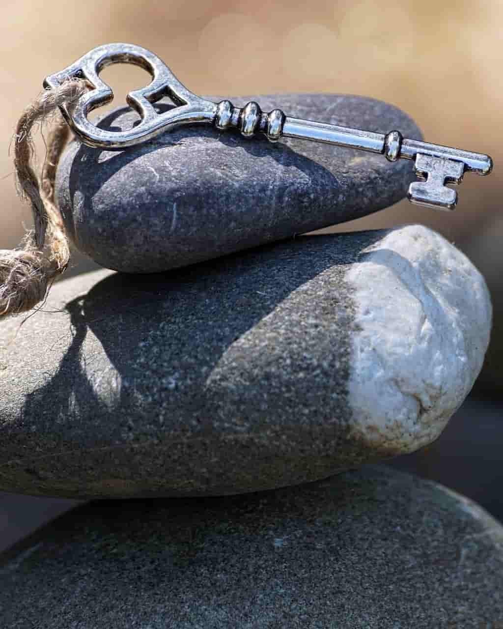 An antique silver-coloured metal key with a natural fibre ribbon resting on the top stone of a stack of three grey stones outdoors.
