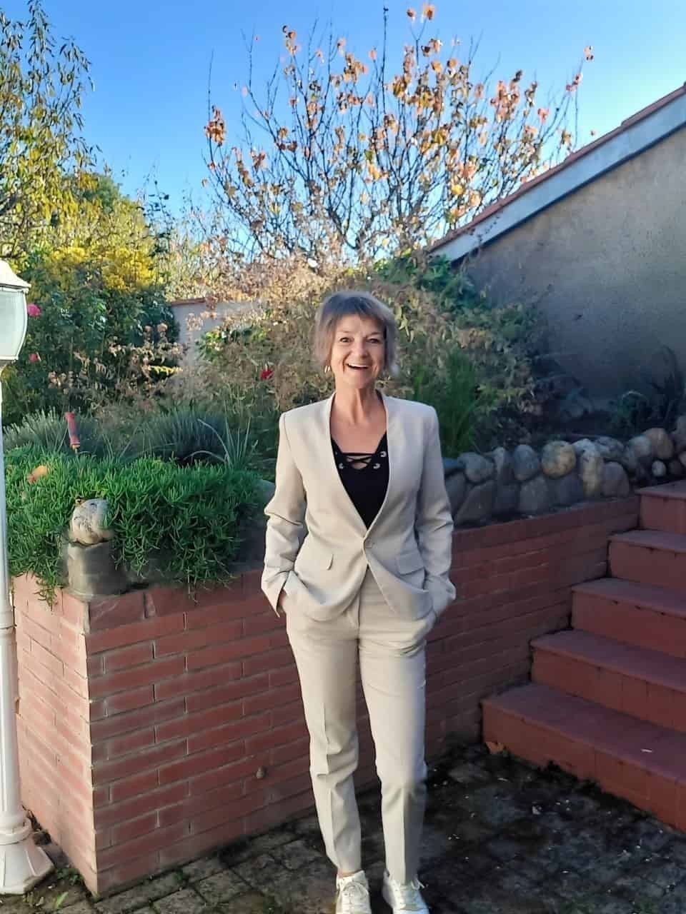 Katja Rehse debout dans son jardin, riant et les mains dans les poches d’un tailleur pantalon beige clair.