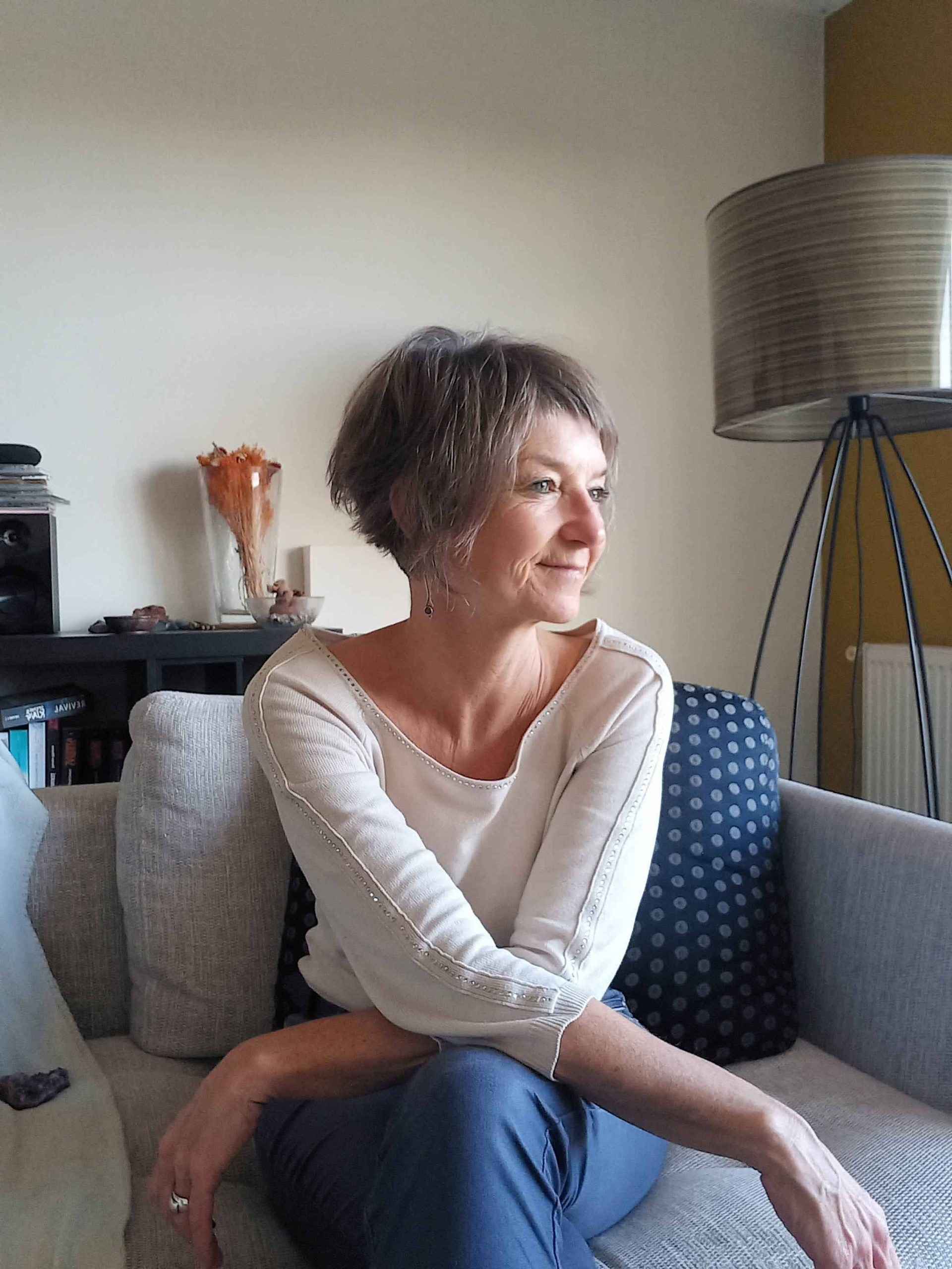 Katja Rehse assise sur un canapé gris clair, les jambes croisées, regardant sur le côté avec un léger sourire.