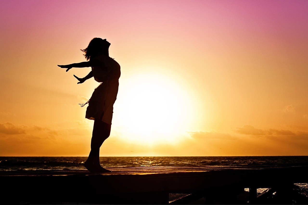 Silhouette of a barefoot woman standing by the water, arms open and face turned towards the sky at sunset.